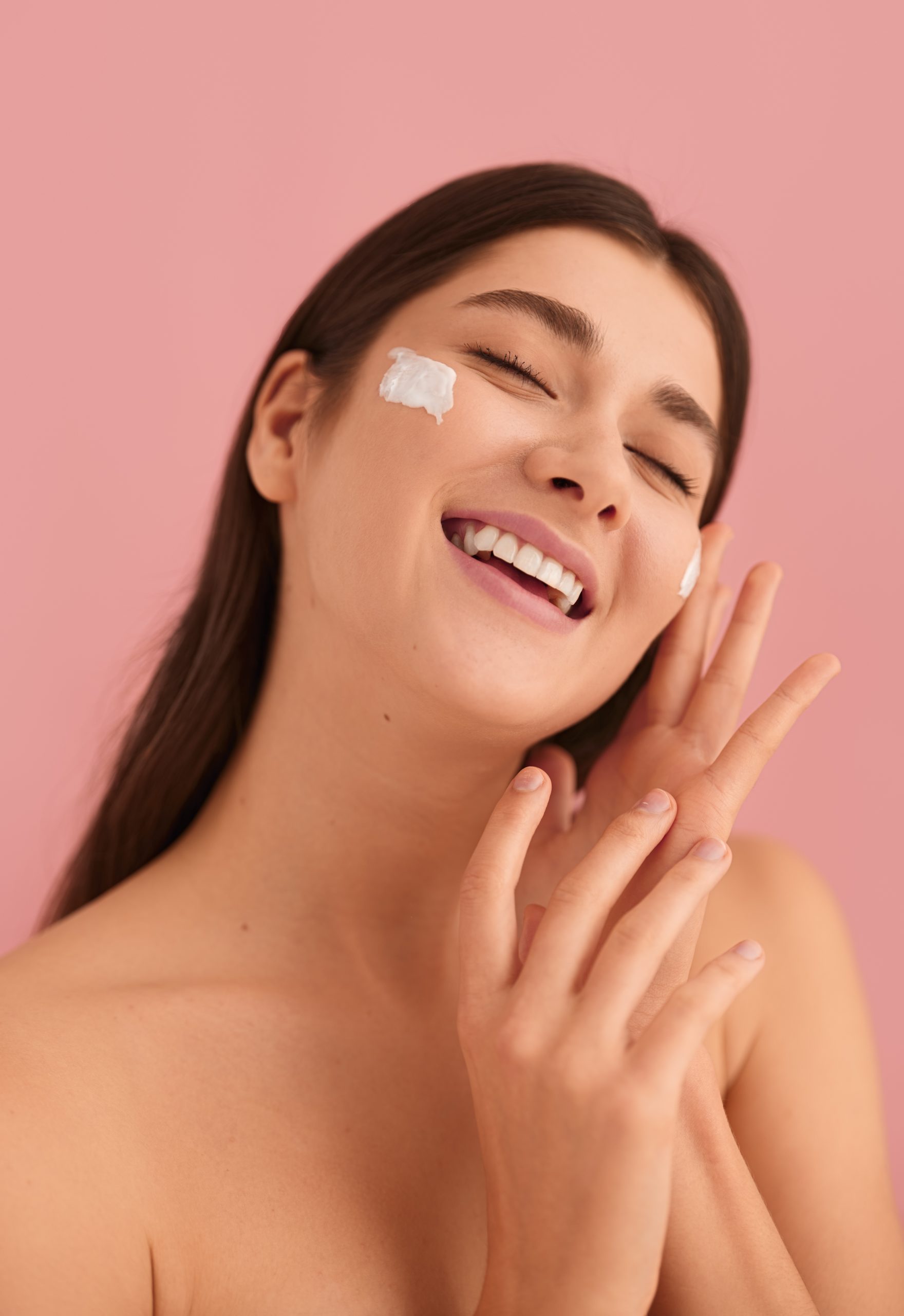 Feminine young woman with perfect skin long dark hair and bare shoulders touching face gently, while applying moisturizing juju cream against pink background during skincare routine